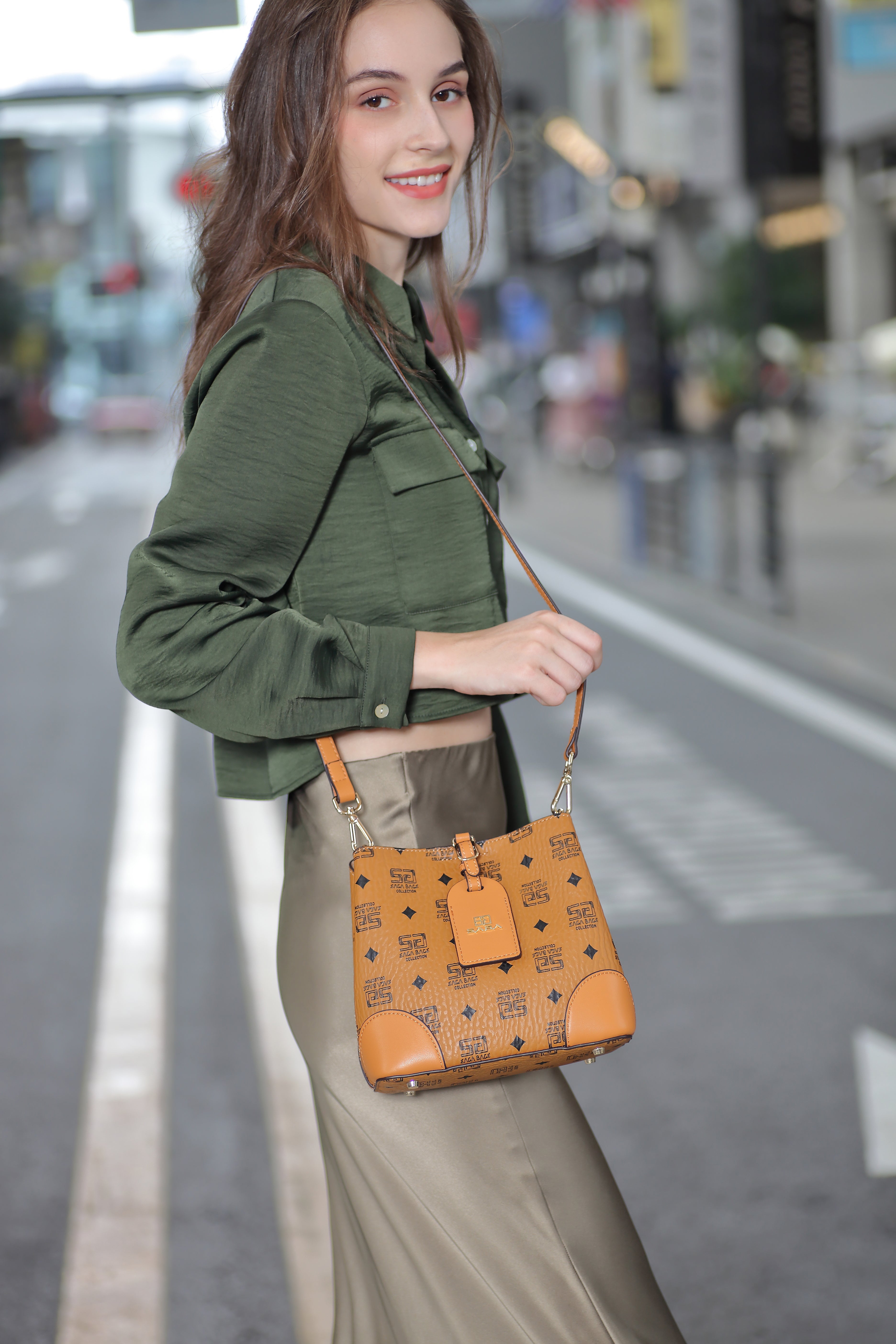 شابة تبتسم وهي تحمل حقيبة يد صغيرة بنية اللون من ماركة ساغا، وترتدي بلوزة خضراء وبنطال بيج في شارع المدينة